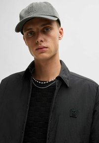 Gray cap with logo, black textured sweater, and black shirt with a subtle shine. Layered silver necklaces with round beads, collar open.