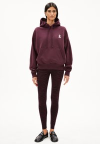 Bordeaux hoodie met een kangoeroe zak en wit lintlogo, gecombineerd met zwart en bordeaux gestreepte leggings. Zwarte instapschoenen.