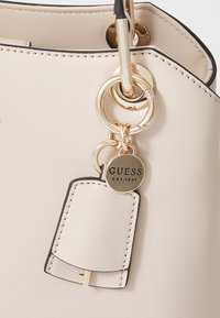 Borsa sintetica beige con hardware dorato, dotata di dettagli con portachiavi e etichetta con logo. Il design include una rifinitura nera.