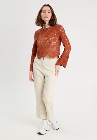 Hæklet langærmet top i rust med blomsterdesign, parret med beige lige ben bukser med et subtilt prikket mønster ved kanten.