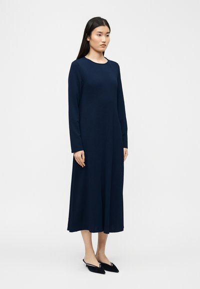 Νεαρή γυναίκα που στέκεται φορώντας μακρυμάνικο μεσοφόρεμα σε ναυτικό μπλε χρώμα και μαύρα παπούτσια χωρίς κορδόνια με μυτερή μύτη, σε απλό ανοιχτό φόντο.