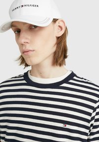 T-shirt di cotone a righe in blu navy e bianco, con scollo a girocollo e un piccolo logo sul petto. Abbinato a un cappellino bianco.