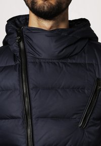 Marineblå pufferjacka med huva, quiltad struktur och två dragkedjefickor fram. Tillverkad av ett slätt, hållbart tyg.