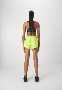 Sujeción deportiva negra y pantalones cortos verde lima brillantes, con cinturilla elástica y corte holgado, combinados con zapatillas deportivas negras.