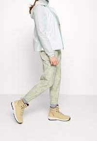 Ljust mintgrön vattentät jacka med grå ärmdetaljer, kombinerad med ljusgröna mönstrade cargo-byxor och beige vandringsskor med svarta accenter.