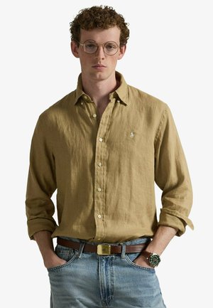 Młody mężczyzna z kręconymi włosami noszący okrągłe okulary, musztardową lnianą koszulę na guziki, niebieskie dżinsy z brązowym paskiem i zegarek na nadgarstku, ręce w kieszeniach.