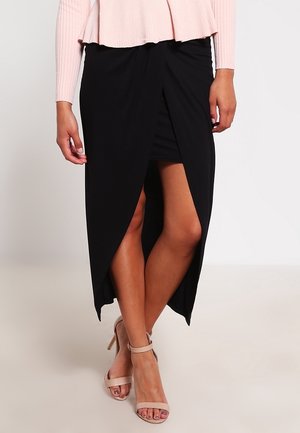 Kobieta ubrana w czarną asymetryczną spódnicę z rozcięciem, jasnoróżowy prążkowany top z długim rękawem oraz beżowe sandały na wysokim obcasie, stojąca ze skrzyżowanymi nogami.