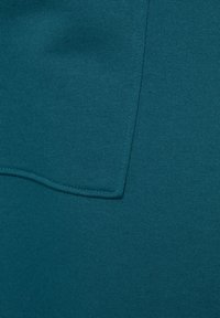 Teal venytyskangas, jossa on sileä rakenne ja hieno ribbaus, varustettuna suurella läpällä vasemmalla puolella. Ei näkyviä kuvioita tai koristeita.