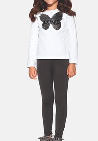 Dziecko ubrane w białą koszulkę z długim rękawem z czarnym motylem i cekinami "LOVE", czarne legginsy oraz szare trampki, stojące na białym tle.