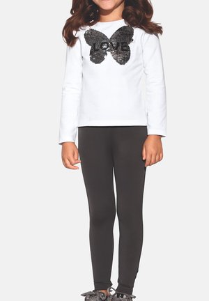 Dziecko ubrane w białą koszulkę z długim rękawem z czarnym motylem i cekinami "LOVE", czarne legginsy oraz szare trampki, stojące na białym tle.