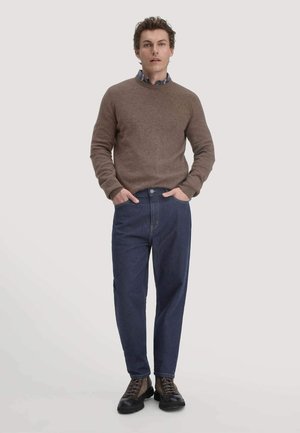 Bruine woltrui, blauwe spijkerbroek en donkere schoenen. De outfit heeft een ronde hals, een relaxte pasvorm en zichtbare zakken op de broek.
