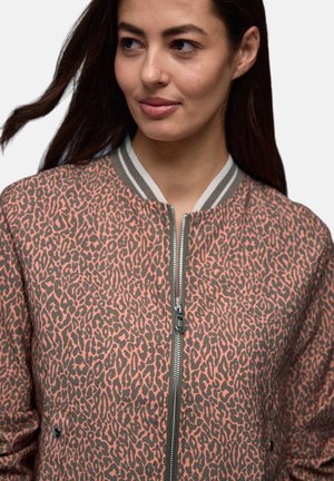 Kobieta z długimi ciemnymi włosami nosi różowo-szarą bluzę na zamek z nadrukiem w cętki i paskowanym kołnierzem, patrząc lekko na bok.