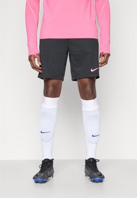 Ροζ μακρυμάνικο μπλουζάκι Nike, μαύρες βερμούδες, λευκές κάλτσες με μωβ λεπτομέρειες και μαύρα παπούτσια ποδοσφαίρου με μπλε σόλες.