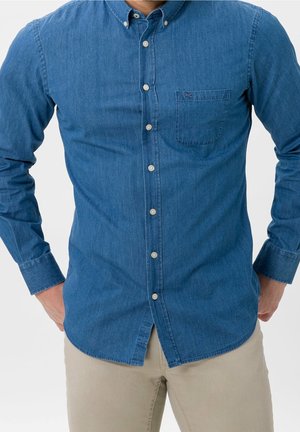 Mand iført blå denim skjorte med knapper og brystlomme samt beige bukser, hænderne hvilende på hofterne mod en ensfarvet baggrund.