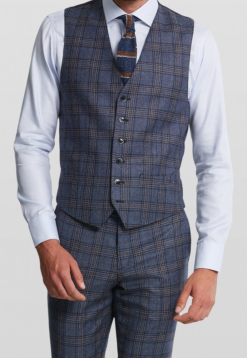 Hombre vistiendo chaleco de cuadros azul y marrón y pantalones a juego con camisa de vestir azul claro y corbata de punto azul con rayas.