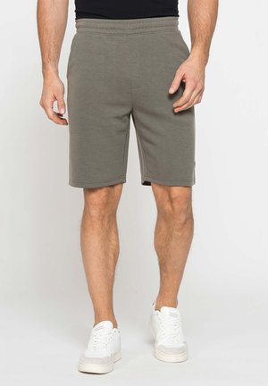 Uomo con pantaloncini verde oliva fino al ginocchio, top nero e sneakers bianche, in piedi su uno sfondo chiaro semplice.