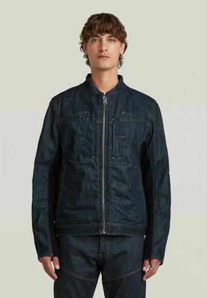 Giovane uomo con capelli ricci corti che indossa una giacca di jeans blu scuro con zip e pantaloni di jeans abbinati, in piedi davanti a uno sfondo semplice.