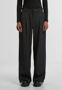Pantaloni cu dungi verticale negre și albe, cu un design cu picioare late, pliuri și un talie ajustată. Purtați cu pantofi masivi negri.