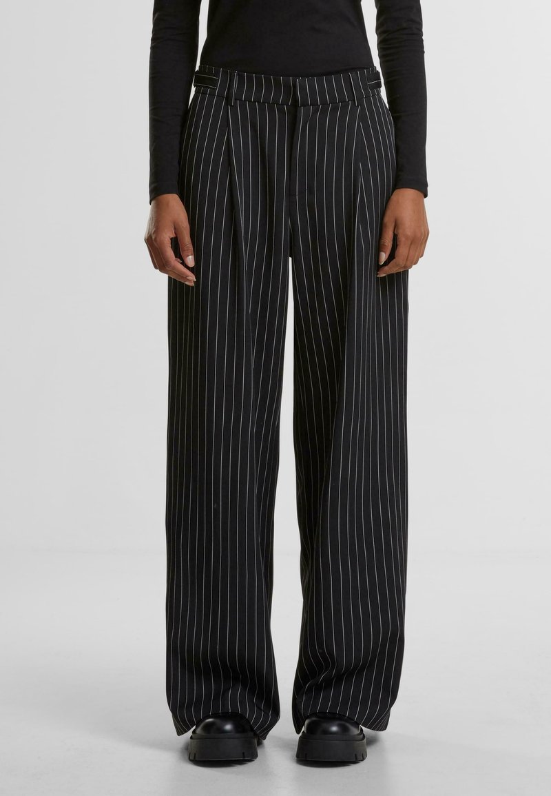 Pantaloni cu dungi verticale negre și albe, cu un design cu picioare late, pliuri și un talie ajustată. Purtați cu pantofi masivi negri.