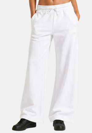 Persona che indossa pantaloni della tuta bianchi larghi con coulisse in vita e scarpe da ginnastica nere, in piedi con le mani in tasca.