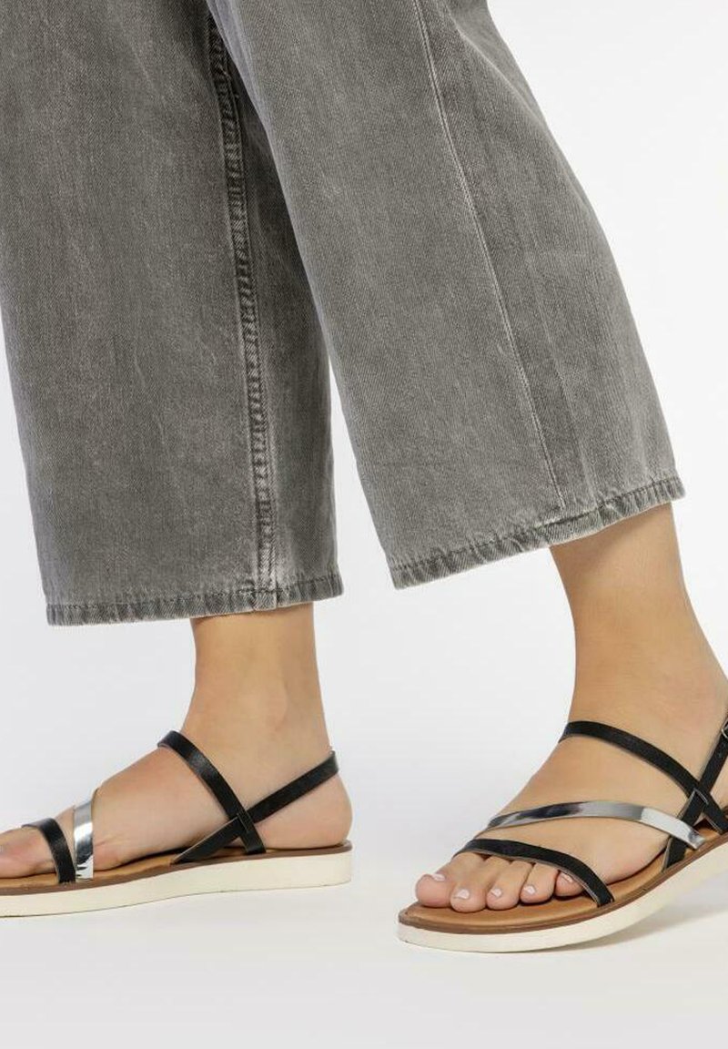 Sandalias negras con tiras y un acento metálico plateado, suela plana, y una mezcla de materiales de cuero y sintético, combinadas con jeans amplios grises.