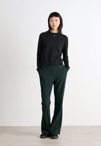 Chemise noire à manches longues avec un col rond, associée à un pantalon vert foncé avec des jambes droites et des poches avant. Des chaussures noires complètent l'ensemble.