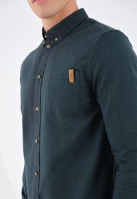 Uomo che indossa una camicia buttoned blu scuro con taschino e etichetta marrone, mostrato dalla gola alla vita su uno sfondo bianco.