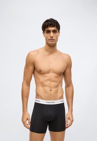 Mladý muž stojící na jednobarevném pozadí, oblečený v černých trenkách s potiskem značky Calvin Klein a bílým pasem.