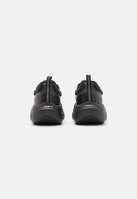 Scarpe da ginnastica nere con tomaia texturizzata, linguetta e suola in gomma con un motivo di battistrada evidente. Design minimalista nel complesso.