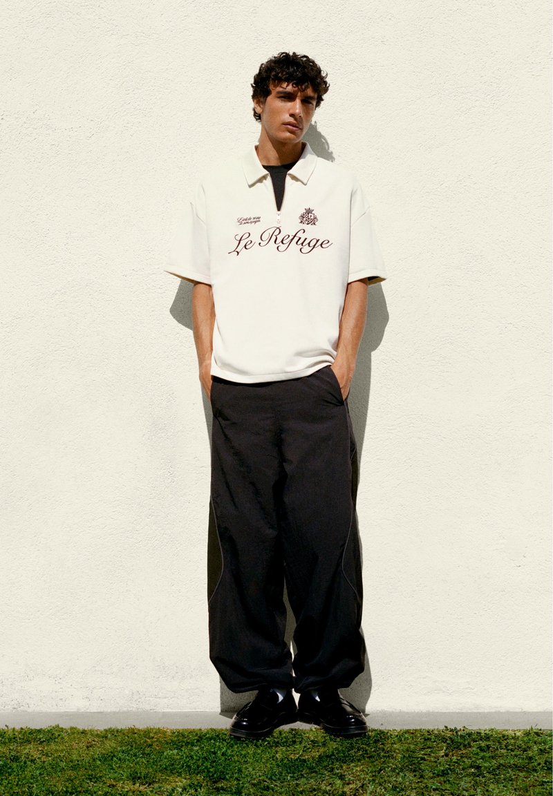 Homme aux cheveux bouclés, debout contre un mur beige, portant un haut blanc à demi-fermeture éclair avec le texte "Le Refuge", pantalon noir et chaussures noires.