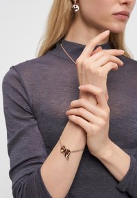 Kvinna som bär en lila långärmad topp med ett rosaguld armband med abstrakta berlocker och matchande rosaguldörhängen.