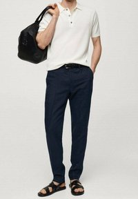 Polo blanc côtelé, pantalon bleu marine foncé, ceinture noire, sac à bandoulière en cuir noir, et sandales noires à larges brides.