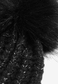 Must värviline kudumiga müts, millel on suur must faux fur pom-pom. Tekstuur sisaldab paksu lõnga ja metallilisi aktsente, mis on läbi kudumisega kombineeritud.