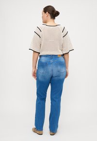 Suéter bege de malha solta com acabamentos pretos, combinado com jeans de denim azul. Os jeans têm corte reto e bolsos traseiros. Calçado castanho completa o visual.