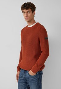 Roestkleurige gebreide trui met geribbelde textuur en ronde hals. Heeft witte accenten bij de kraag en een logopatch op de linkerarm.