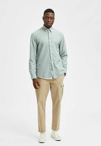 Lichtgroen overhemd met knoopsluiting en kraag, gecombineerd met beige cargobroek en witte sneakers. Het overhemd heeft een zachte, gestructureerde stof.
