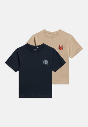 To kortermede t-skjorter med rund hals, en beige med en rød krabbelapp, en marineblå med en blå fiskelapp, lagt flatt på en hvit bakgrunn.