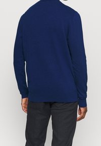 Marinblå stickad tröja med lång ärm, rundad halsringning och ribbat avslut. Slät yta med en figurnära design, bärs med grå byxor.