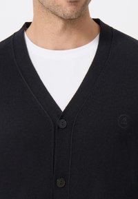 Černý pletený cardigan s výstřihem do V, který má tři knoflíky a jemný logotyp na levé hrudi. Měkká textura.