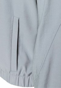 Light grey windbreaker jacket with a smooth texture, elastic hem, and side pocket. Features minimalist design and flat seams for durability.