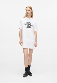 Bijela haljina majica kratkih rukava s crnim grafičkim natpisom "KARL LAGERFELD JEANS", opuštenog kroja, od glatkog pamučnog materijala i nejednakim obrubom. Crne cipele i čarape.