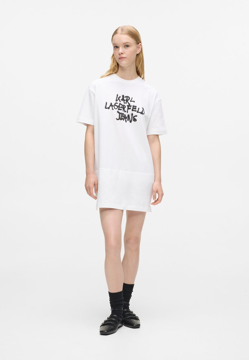 Bijela haljina majica kratkih rukava s crnim grafičkim natpisom "KARL LAGERFELD JEANS", opuštenog kroja, od glatkog pamučnog materijala i nejednakim obrubom. Crne cipele i čarape.