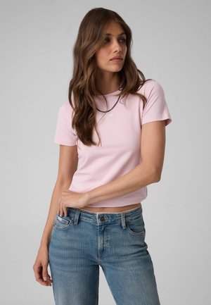 Frau mit langen braunen Haaren, die ein hellrosa kurzärmliges Shirt und blaue Jeans trägt, steht mit einer Hand an der Taille vor einem schlichten Hintergrund.