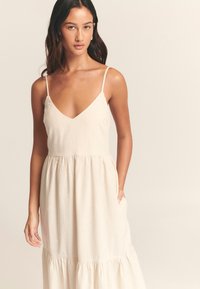 Light beige sleeveless dress with a deep V-neck, gathered waist, and tiered ruffled hem. Fabric has a soft, textured appearance.