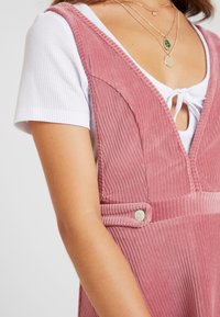 Roze corduroy jurk met een diepe V-hals, textiel met structuur en zijknopdetail, gelaagd over een witte aansluitende blouse.