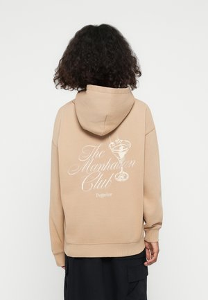 Beige hoodie met een witte grafische print van een cocktailglas en de tekst "The Manhattan Club Pegador" op de achterkant.