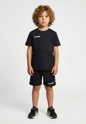 Dziecko z kręconymi włosami, ubrane w czarną odzież sportową Hummel, w tym koszulkę, szorty i trampki, stojące na jednolitym tle.