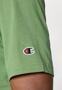 Grünes Baumwoll-T-Shirt mit kurzen Ärmeln, das auf dem linken Ärmel ein kleines gesticktes Logo in Rot und Schwarz zeigt. Glatte Textur.