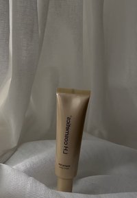 Tube rouge en métal doré avec un design texturé, étiqueté "LH Cosmetics", comportant un texte noir et un capuchon en relief, posé sur un tissu blanc.
