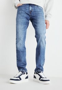 Vaqueros de denim azul claro con un corte clásico, que presentan un desvanecimiento sutil. Combinados con zapatos blancos acentuados con detalles en azul marino y herrajes en negro.